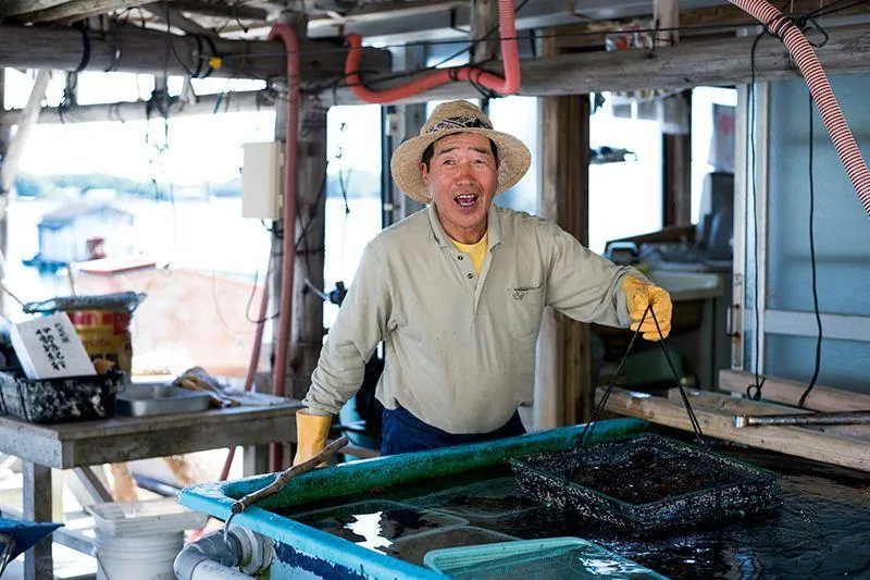 Japanese pearl farmer holding oyster baskets beside a coastal workshop